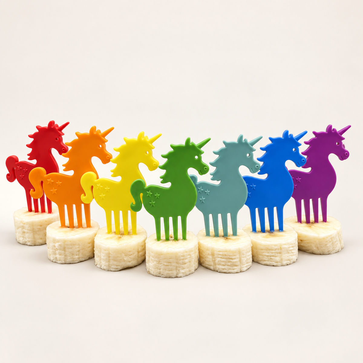 Sieben bunte Pieksi Snack-Piekser in Einhornform aus dem Set „Cuddle Corns“, aufgesteckt auf Bananenscheiben.