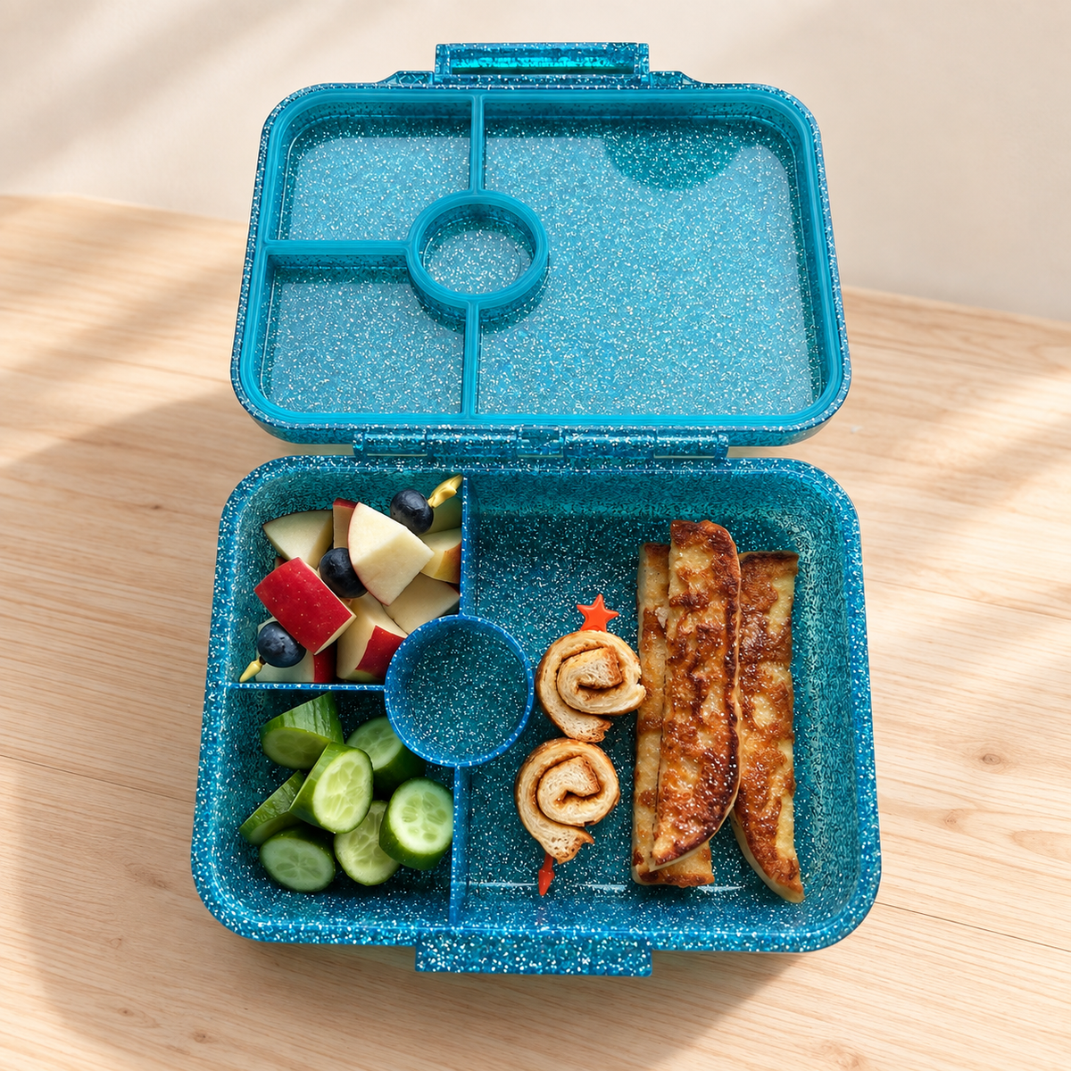 Geöffnete LEKKABOX Glamour Glitzer-Brotdose in Aqua mit vier Fächern, gefüllt mit Obst, Gurken und Gebäck.