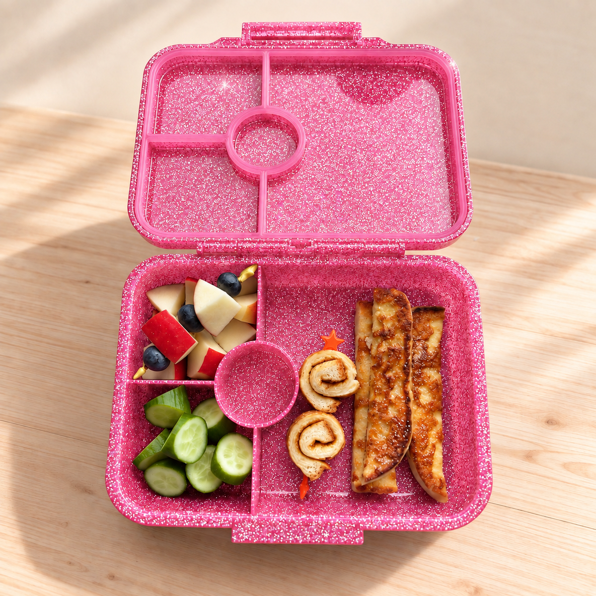 Geöffnete LEKKABOX Glamour Glitzer-Brotdose in Pink mit vier Fächern, gefüllt mit Obst, Gurken und Gebäck.
