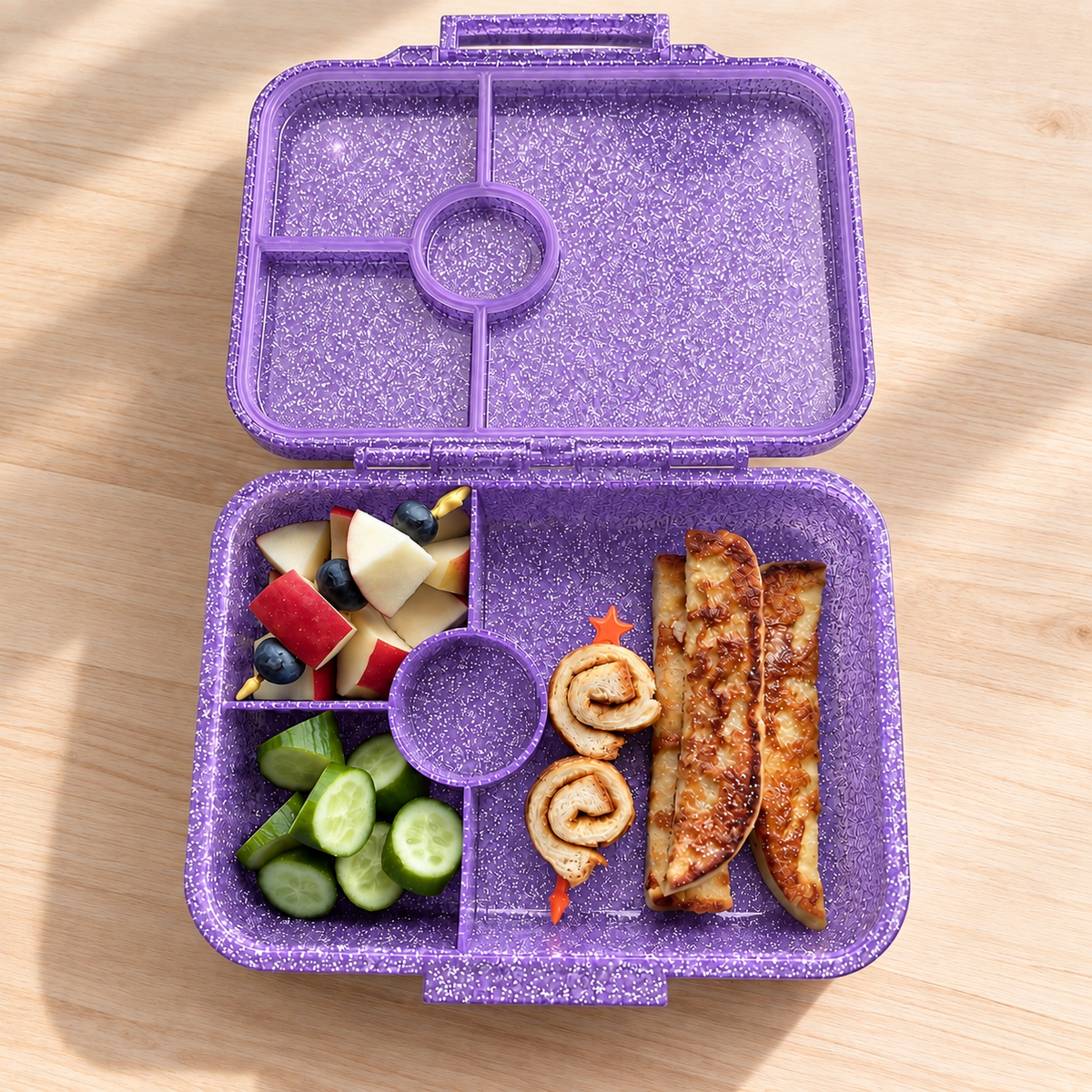 Geöffnete LEKKABOX Glamour Glitzer-Brotdose in Purple mit vier Fächern, gefüllt mit Obst, Gurken und Gebäck.